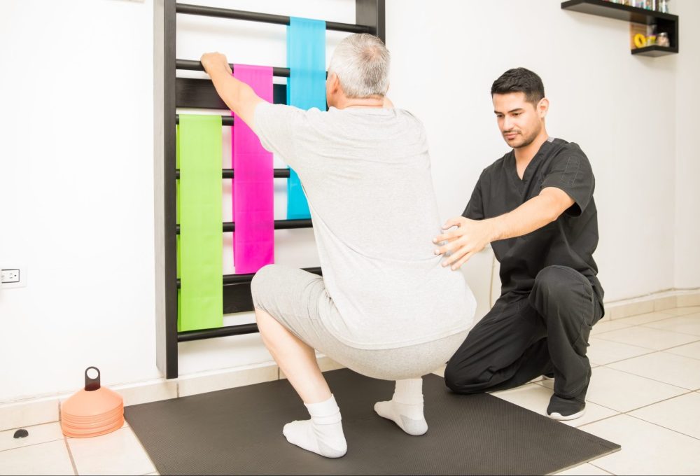 Therapist Assisting Mature Man Doing Squats In Clinic